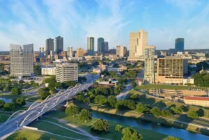 Aerial view of downtown cityscape with modern skyscrapers, river, green spaces, and bridge infrastructure.