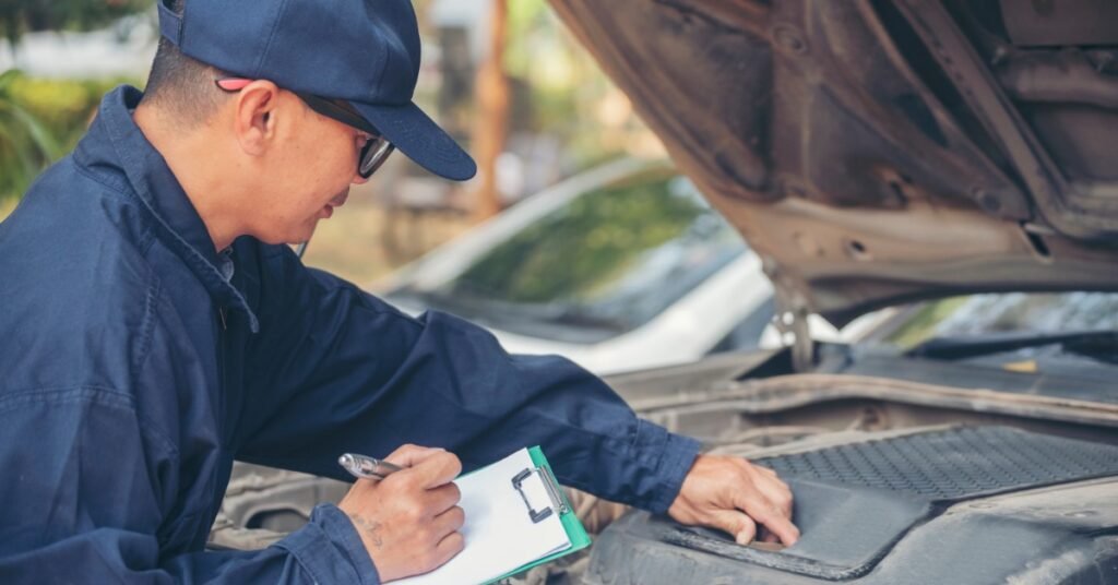 A close-up of a person in a blue uniform writing on a clipboard while checking under the raised hood of a dusty car.