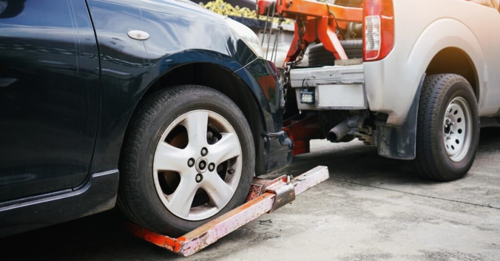 A black car is being loaded onto a white tow truck with a red metal lift beneath the front tire on a concrete roadside.