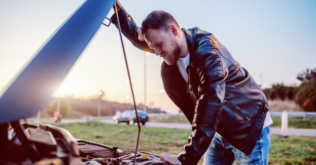 A close-up of a man in a leather jacket looking at a car engine under an open hood, on the side of a road.
