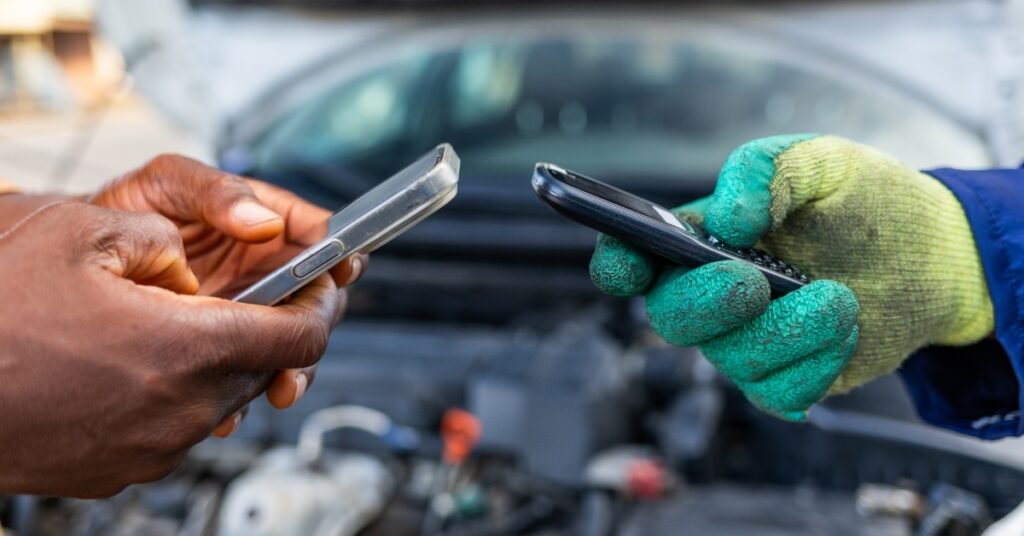 A close-up of two people holding smartphones in front of an open car hood with a blurred view of the engine.