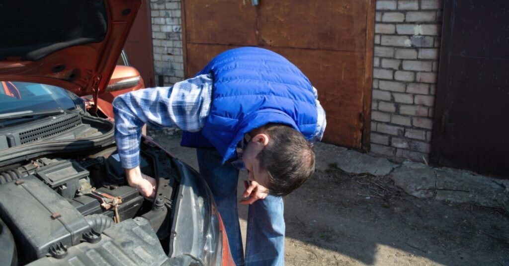 A close-up of a man in a blue vest leaning over the engine of an orange car with its hood open outside a brick garage.
