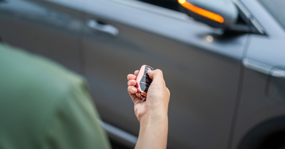 A hand holding a black car key fob with a thumb on the buttons, beside a gray vehicle with an illuminated side mirror.