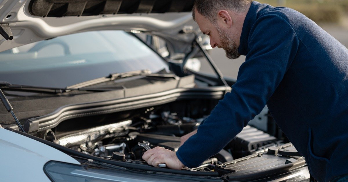 A man wearing a blue jacket leans over the open hood of a silver car and examines components inside the engine bay.