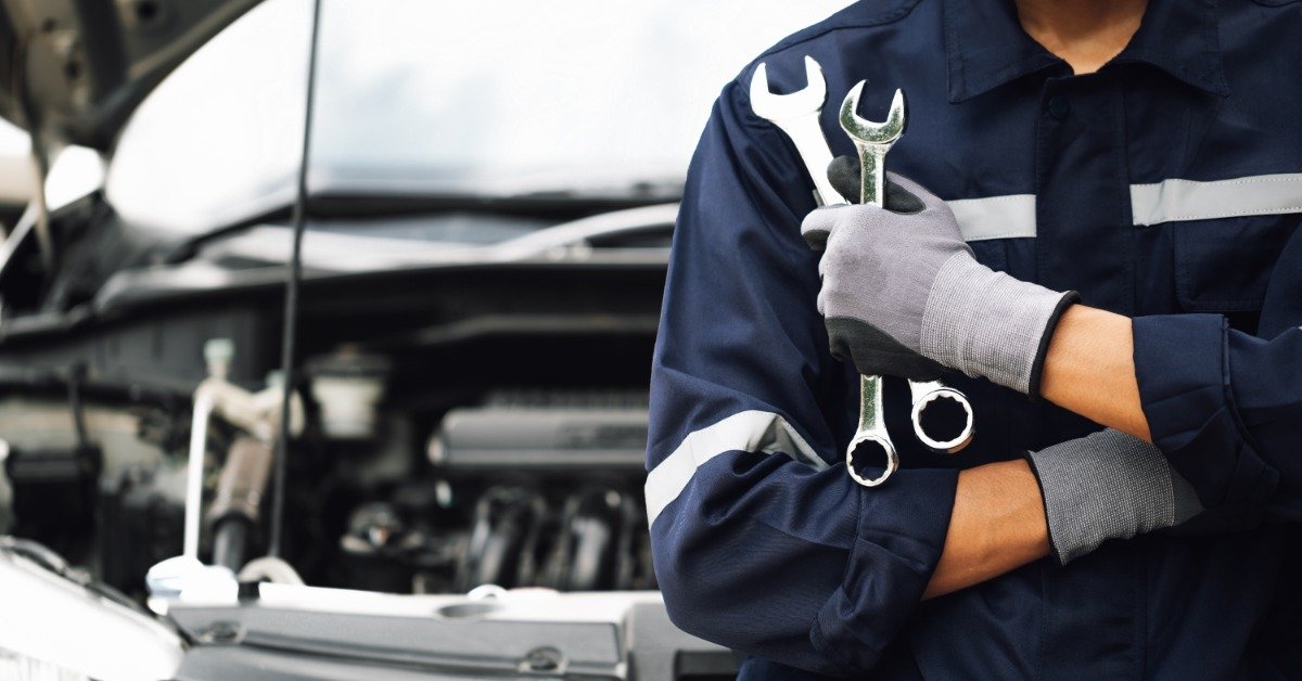 A person in a work uniform and gray gloves holds two metal wrenches beside a car with its hood raised.